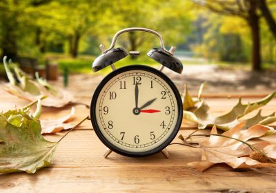 Daylight Saving Time, Fall Back one hour. Black alarm clock with time change on wooden table. Autumn trees and leaves background