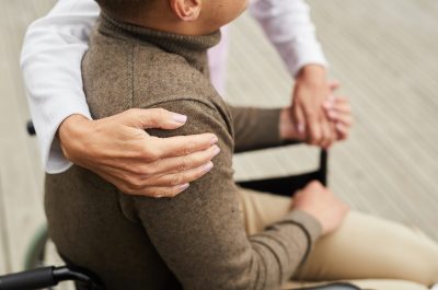 Close-up of unrecognizable medical specialist embracing shoulder of patient while helping him to stand up out of wheelchair